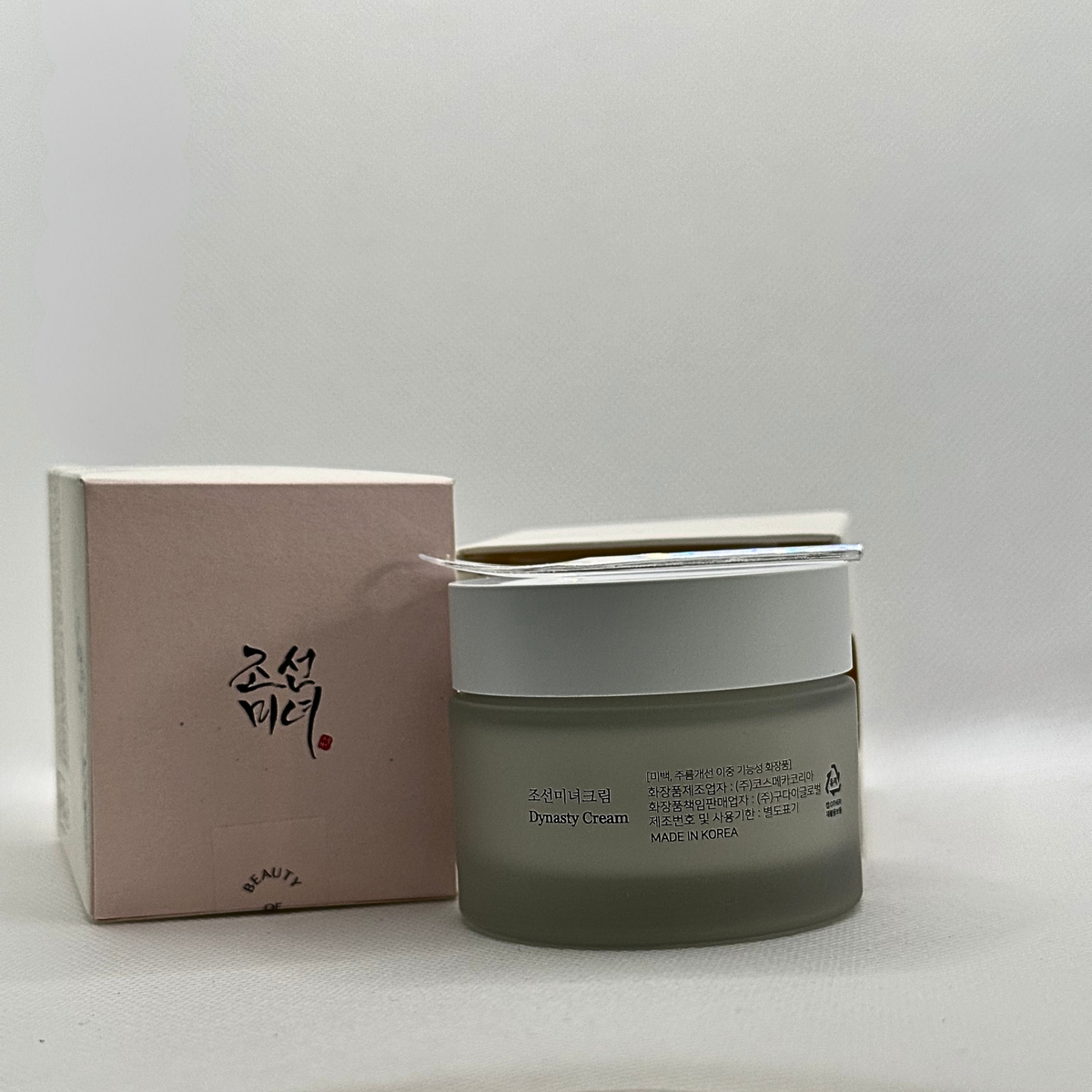 A jar of Dynasty Cream with a frosted gray-green glass body and a white lid, shown next to its light pink packaging box on a neutral light gray background. Display is of the back of the jar, next to its box with the plastic spatular resting on top of the jar.