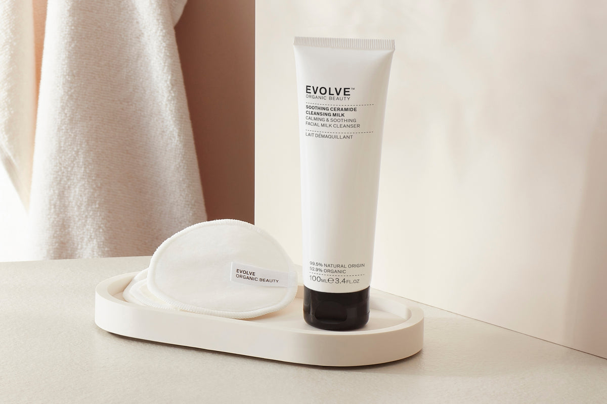 Evolve Soothing Ceramide Cleansing Milk in a white squeeze tube with a black cap, displayed next to a white cotton pad on a round cream tray, with a soft beige towel in the background.