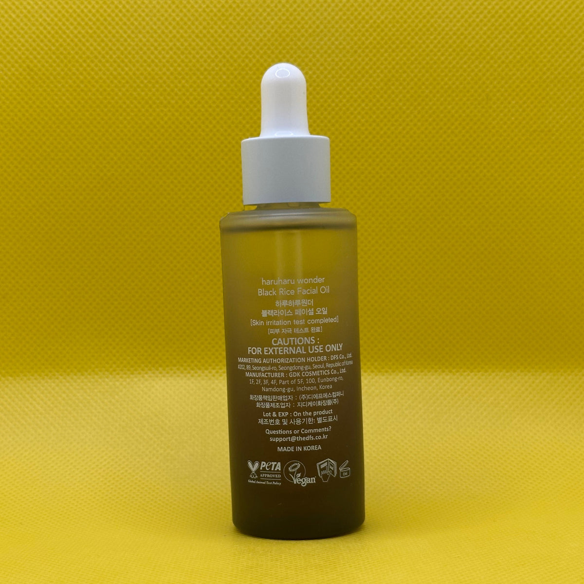 The back of a bottle of Haruharu Wonder Black Rice Facial Oil, featuring a white dropper cap and a dark amber-brown bottle, set against a bright yellow textured background.
