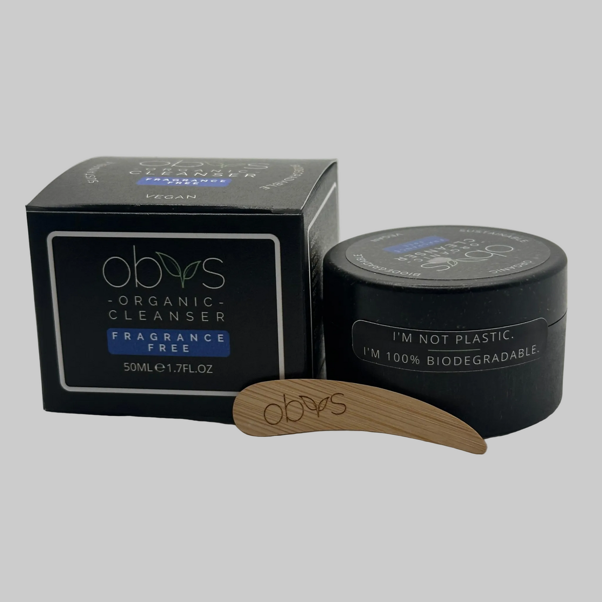 A black box and black jar of obvs Organic Cleanser next to a small wooden applicator with the obvs logo, on a light gray background. The jar has a sticker stating: I'm not plastic. I'm 100% Biodegradable.