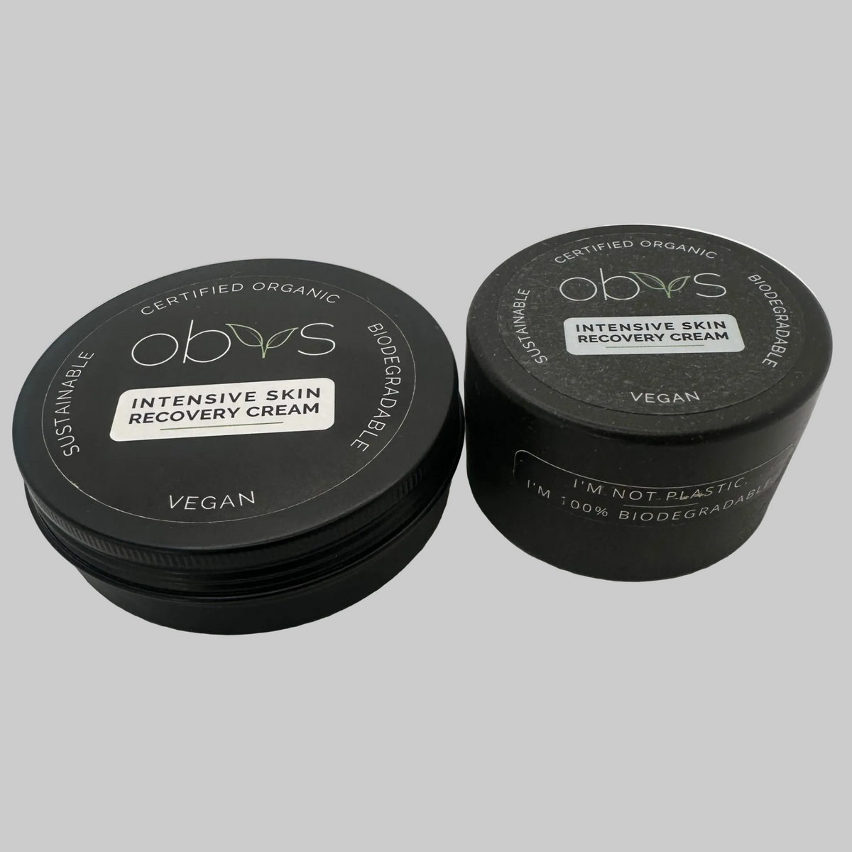 A 100ml tin of Obvs Intensive Recovery Cream next to a biodegradable jar of Obvs Intensive Skin Recovery Cream (50ml). The labels read: Certified Organic, Biodegradable, Vegan, Sustainable. Obvs Intensive Recovery Cream.