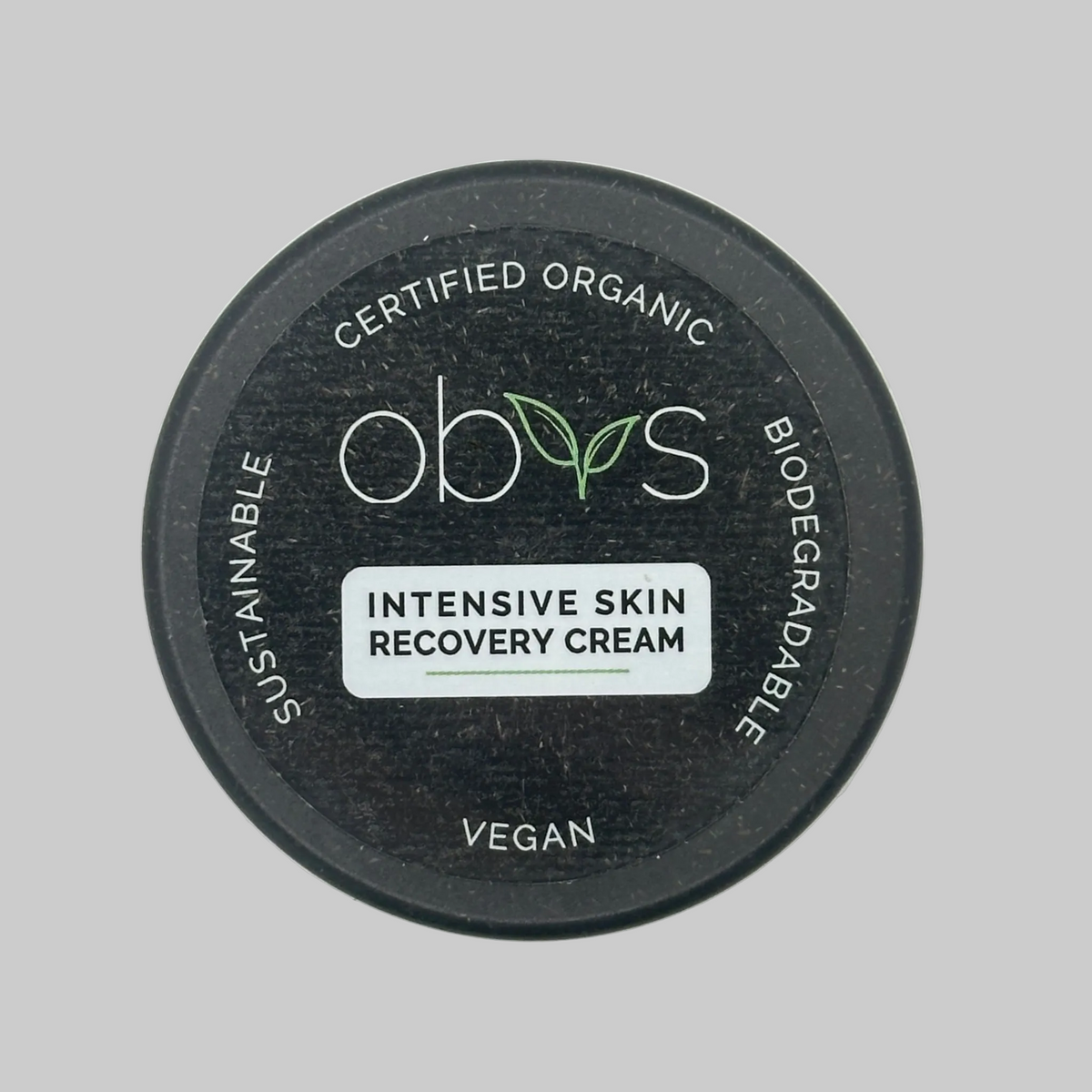 Top-down view of a round black biodegradable jar lid for Obvs Intensive Skin Recovery Cream, featuring white and green text and a white label, on a light grey background. Label reads: Certified Organic, Biodegradable, Vegan, Sustainable. Obvs Intensive Recovery Cream.