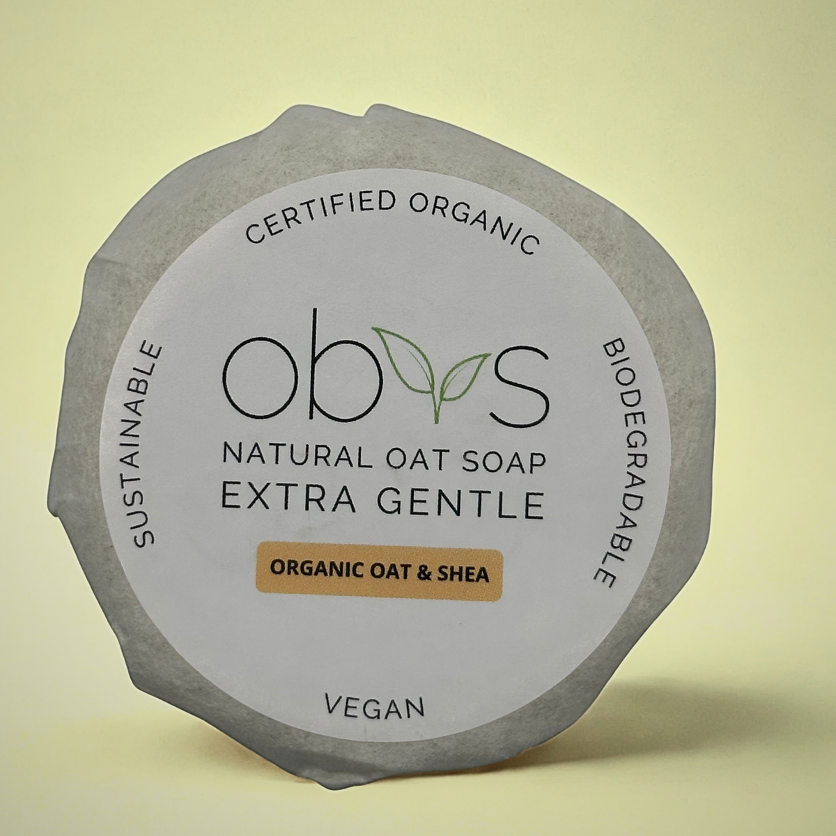 Obvs Natural Oat Soap, wrapped in tissue shown on a plain light yellow background. Writing on the round white label state: Certified Organic, Biodegradable, Vegan, Sustainable. Obvs natural oat soap extra gentle. Organic oat and shea.