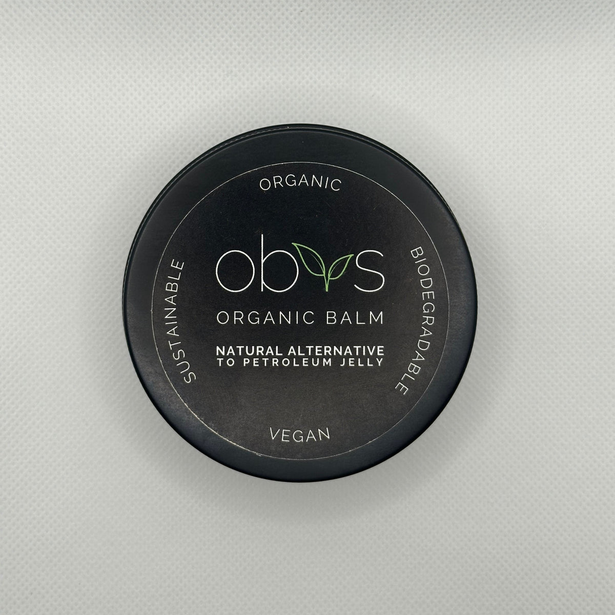 A round black tin of Obvs Organic Balm with a black lid, featuring white and green text and logo, set against a light gray textured background.
