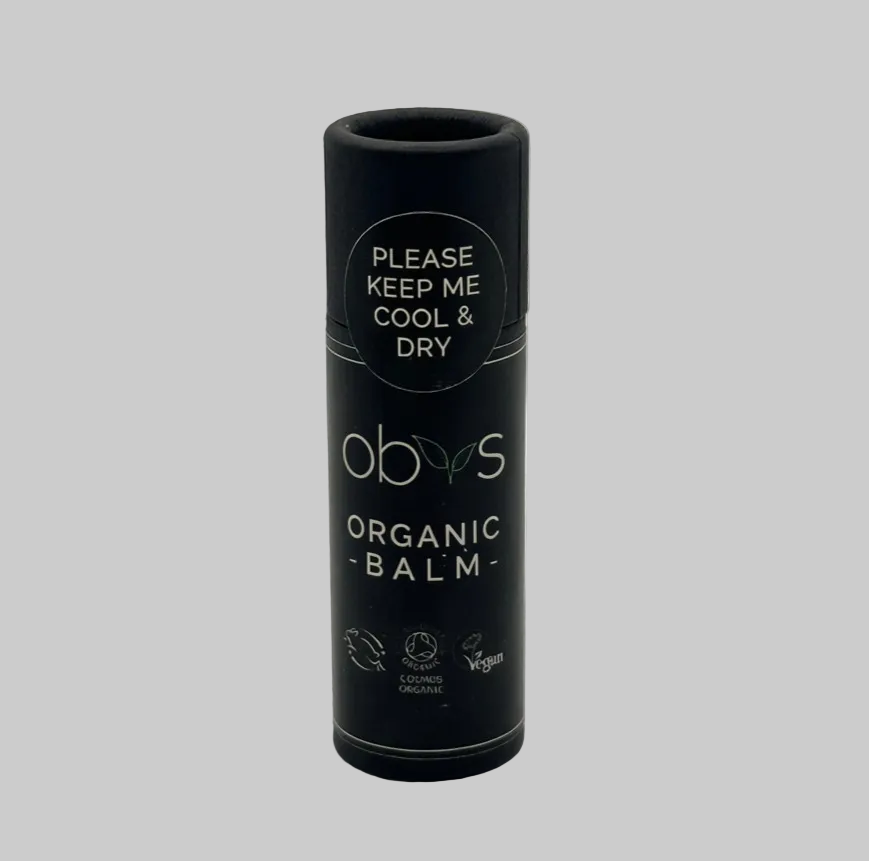 A black cylindrical tube of obvs Organic Balm with white text, photographed against a neutral light gray background. Text reads: Obvs organic balm. Please keep me cool and dry. Certifications for Leaping bunny, COSMOS Organic and Vegan are displayed on the tube.