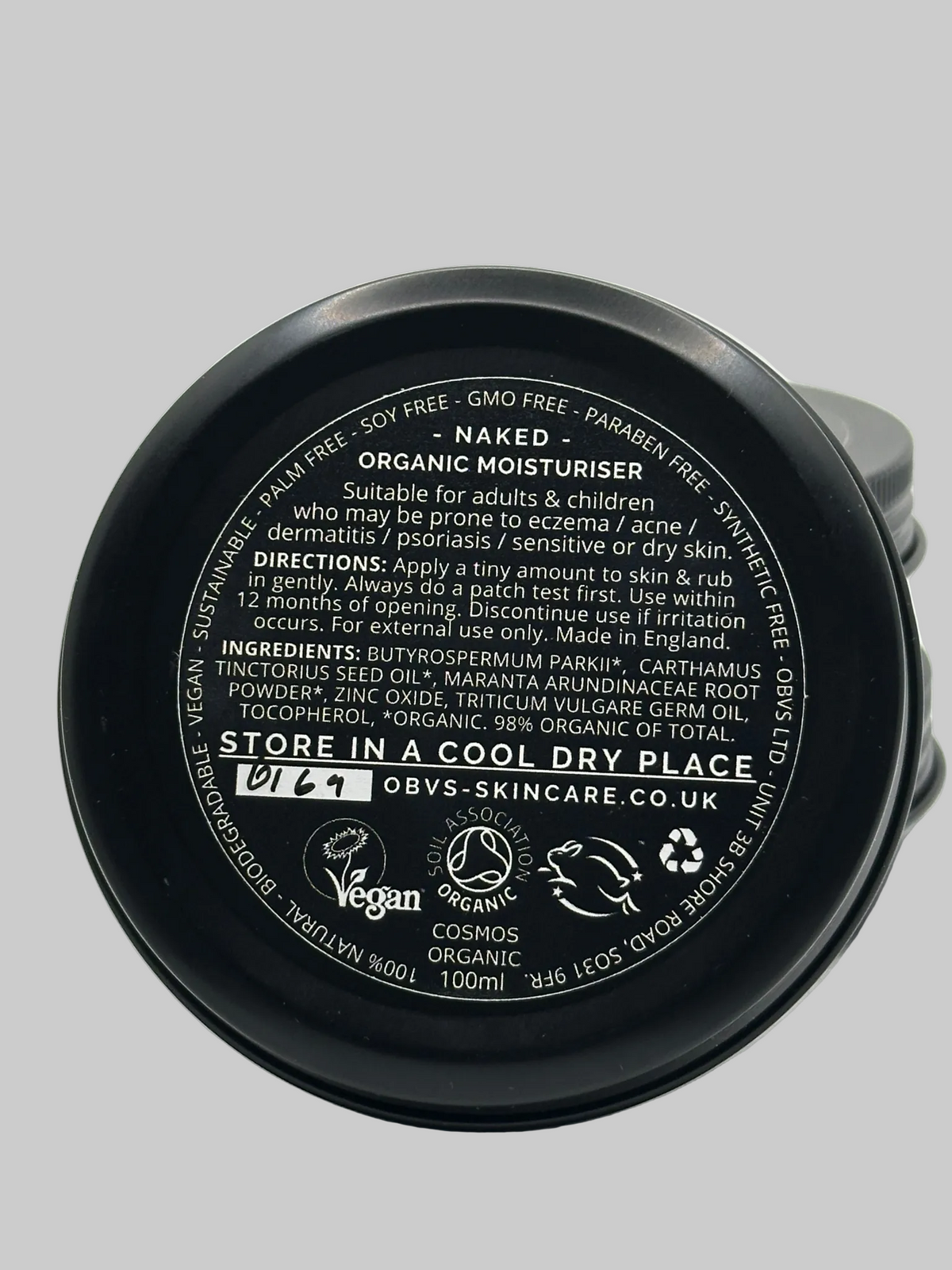 The back label of an Obvs 'Naked' Organic Moisturiser tin, displaying product details, ingredients, and usage instructions on a light gray background. Label states that it is suitable for adults & children who may be prone to eczema/acne/dermatitis/sensitive or dry skin. Made in England. Vegan, COSMOS Organic, Leaping Bunny.