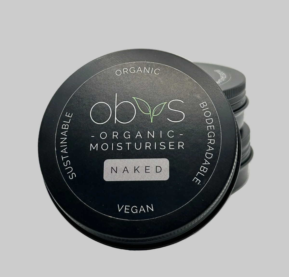 A round black tin of obvs Organic Moisturiser in the Naked (no fragrance), with three more tins stacked behind it on a light gray background. The label reads: Organic, Biodegradable, Vegan, Sustainable. Obvs Organic Moisturiser - Naked.