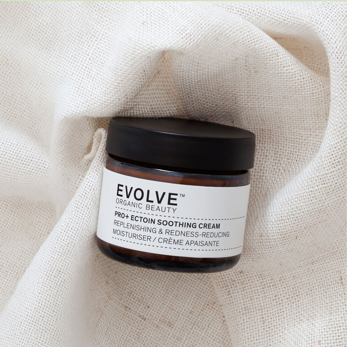 A jar of EVOLVE Organic Beauty Pro+ Ectoin Soothing Cream resting on a textured, draped beige linen fabric background.