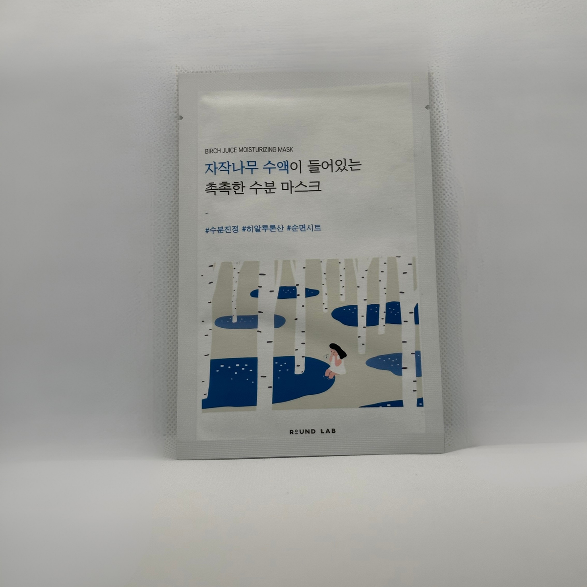A single sheet Round Lab Birch Juice mask packet standing upright on a light gray surface against a white background. The packet is white with blue Korean text and an illustration of birch trees and water at the bottom.
