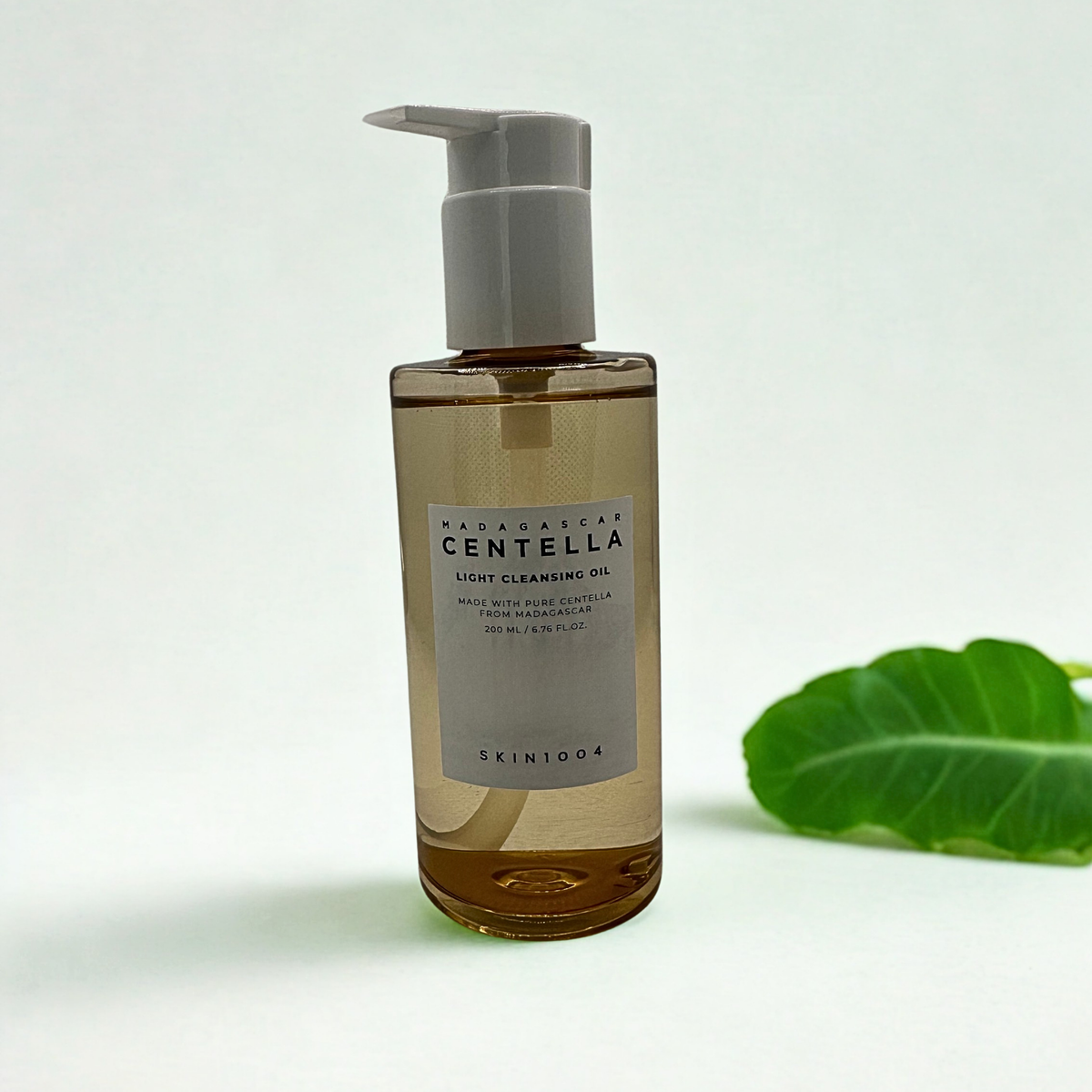 A bottle of SKIN1004 Madagascar Centella Light Cleansing Oil with a white pump dispenser, set against a clean white background with a green leaf on the right. The bottle reads: Madagascar Centella light cleansing oil. Made with pure centella from Madagascar. 200ml
