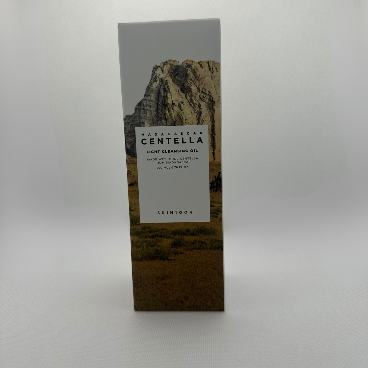 A tall rectangular product box for SKIN1004 Madagascar Centella Light Cleansing Oil, featuring a scenic image of a rocky mountain range on a beige and brown background with a white label in the center.
