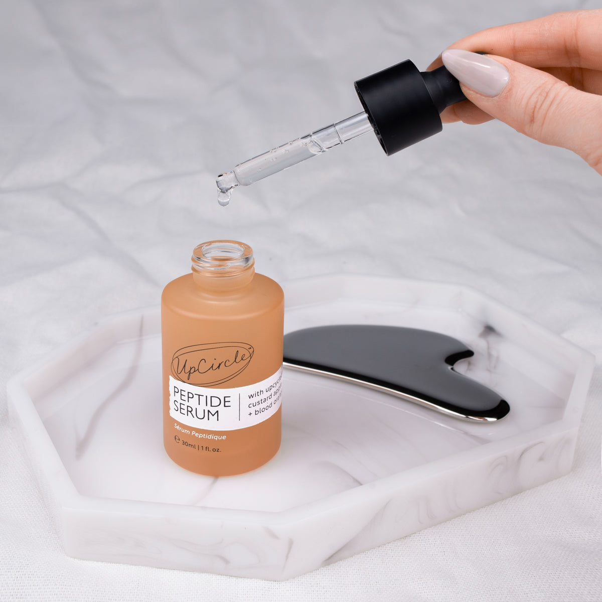 A hand with pale pink manicured nails holds a glass dropper above a bottle of UpCircle Peptide Serum. The bottle is a warm peachy-orange color with a black dropper cap. The serum is a clear colour.