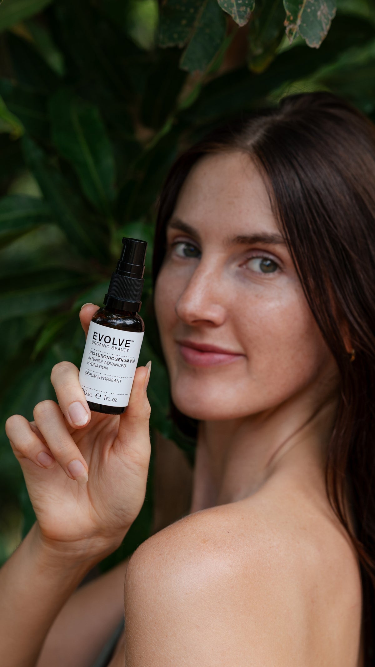 A woman with long brown hair holds up a Evolve Hyaluronic Serum 200 30ml bottle against a background of lush green leaves.