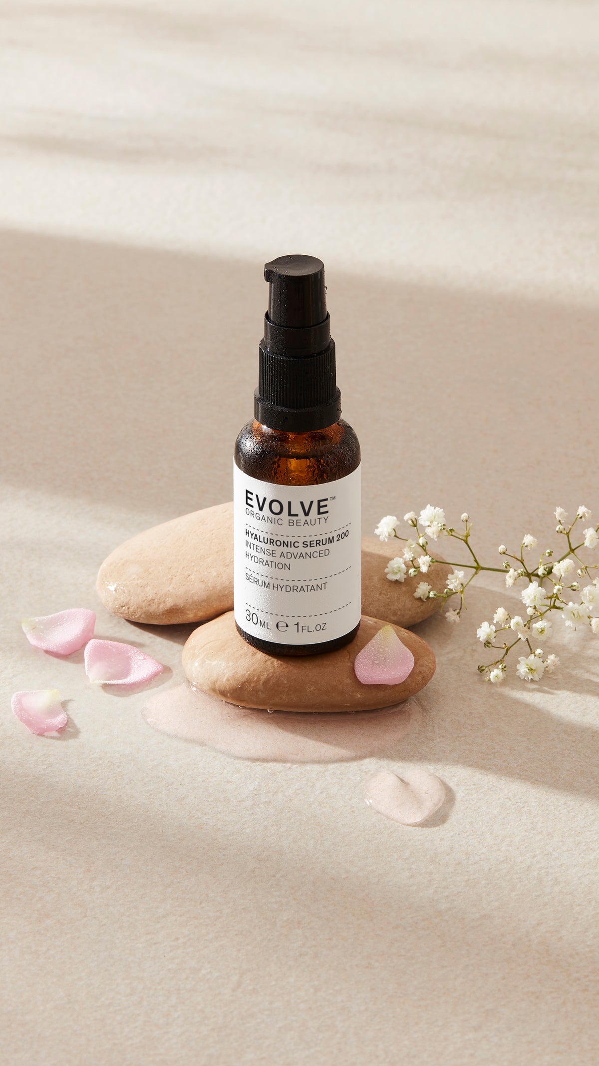 Evolve Hyaluronic Serum 200 30ml bottle with amber glass and a black pump and white label, styled on beige stones with pink petals and baby's breath flowers.