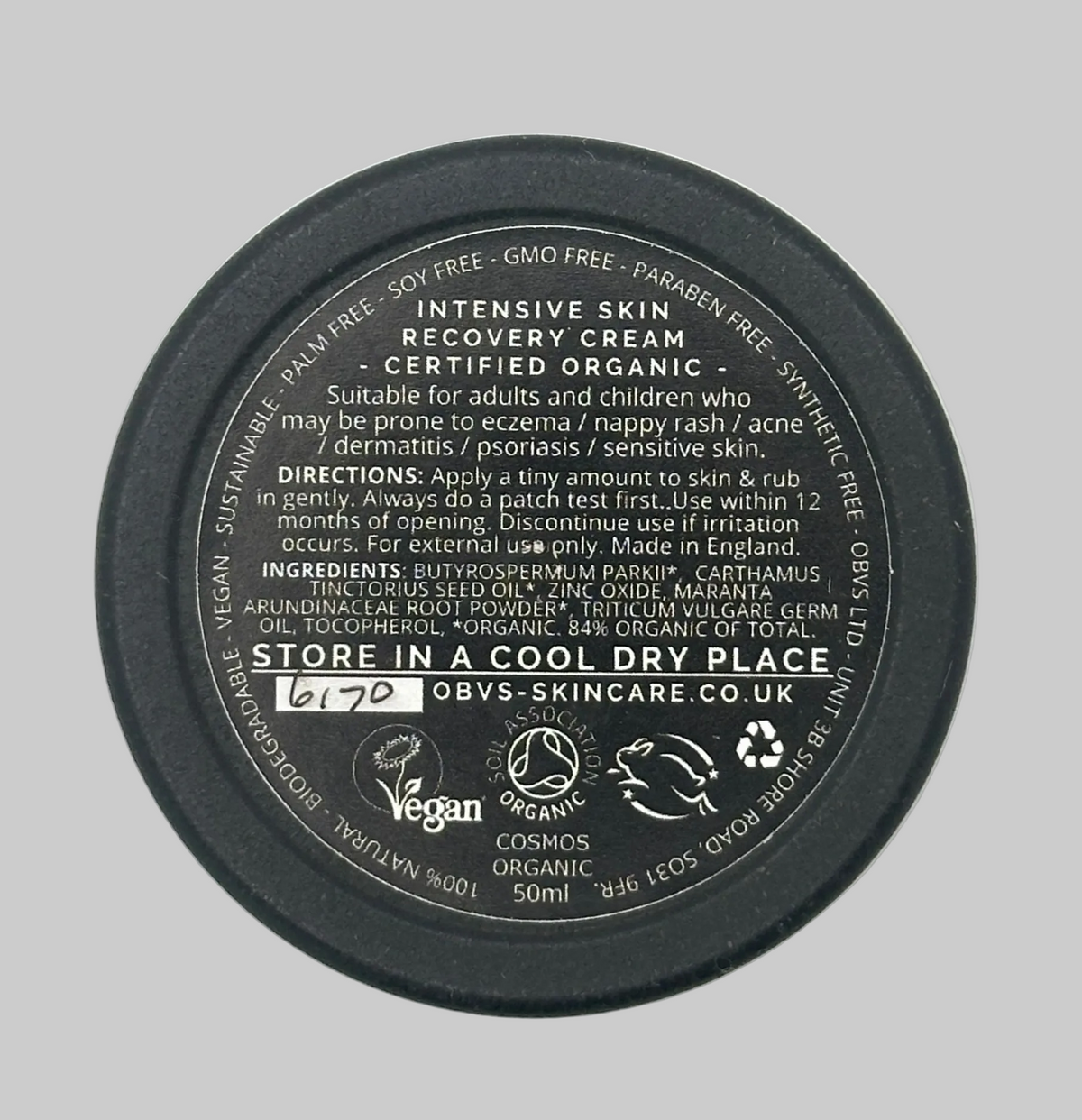 The bottom of a round black jar for OBVS-SKINCARE Intensive Skin Recovery Cream, showing product details, ingredients, and application instructions on a light textured background. Label states that it is suitable for adults and children who may be prone to eczema / nappy rash / acne / dermatitis / psoriasis / sensitive skin. Vegan, COSMOS Organic, Leaping bunny.