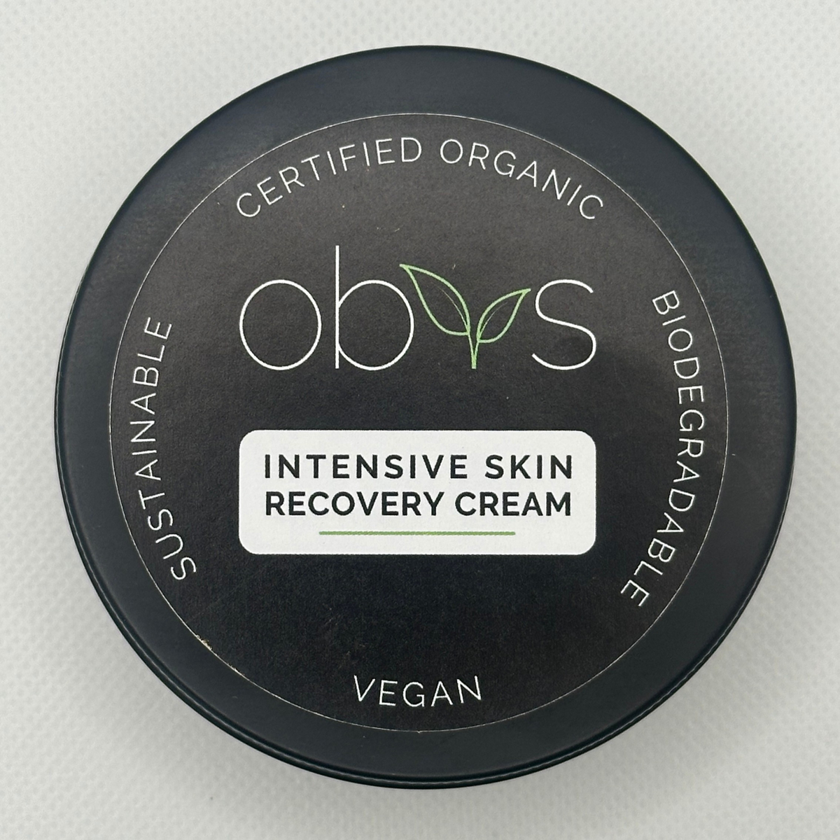 Top-down view of a round black tin lid for Obvs Intensive Skin Recovery Cream, featuring white and green text and a white label, on a light textured background. Label reads: Certified Organic, Biodegradable, Vegan, Sustainable. Obvs Intensive Recovery Cream.