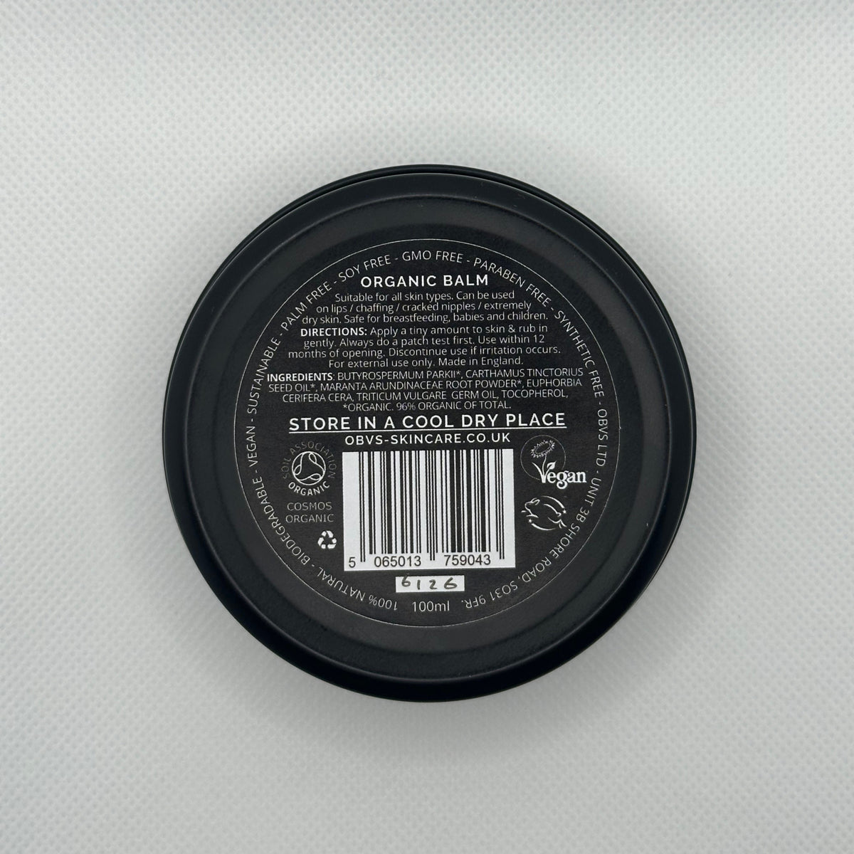 The bottom of a round black cosmetic tin showing the label for OBVS SKINCARE ORGANIC BALM, photographed from above on a light gray textured background.