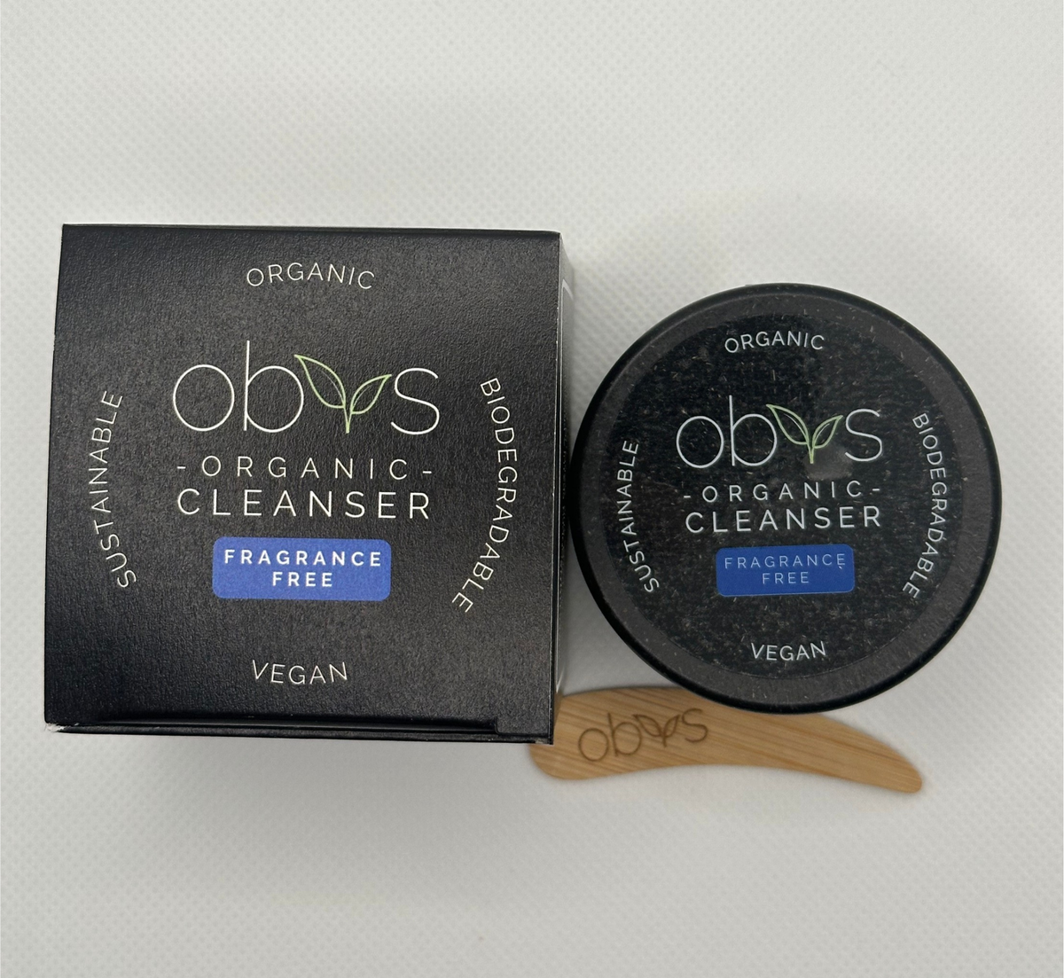 A black square box and round jar of obvs Organic Cleanser with a small wooden spatula, displayed on a light gray textured background. Packaging reads: Organic, Biodegradable, Vegan, Sustainable. Obvs Organic Cleanser. Fragrance Free.