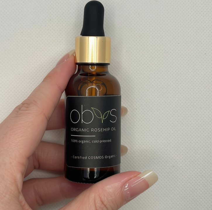 A hand holding 30ml amber glass dropper bottle of obvs Organic Rosehip Oil against a light textured background. Th label reads: Obvs Organic Rosehip Oil. 100% organic, cold pressed. Certified COSMOS organic.
