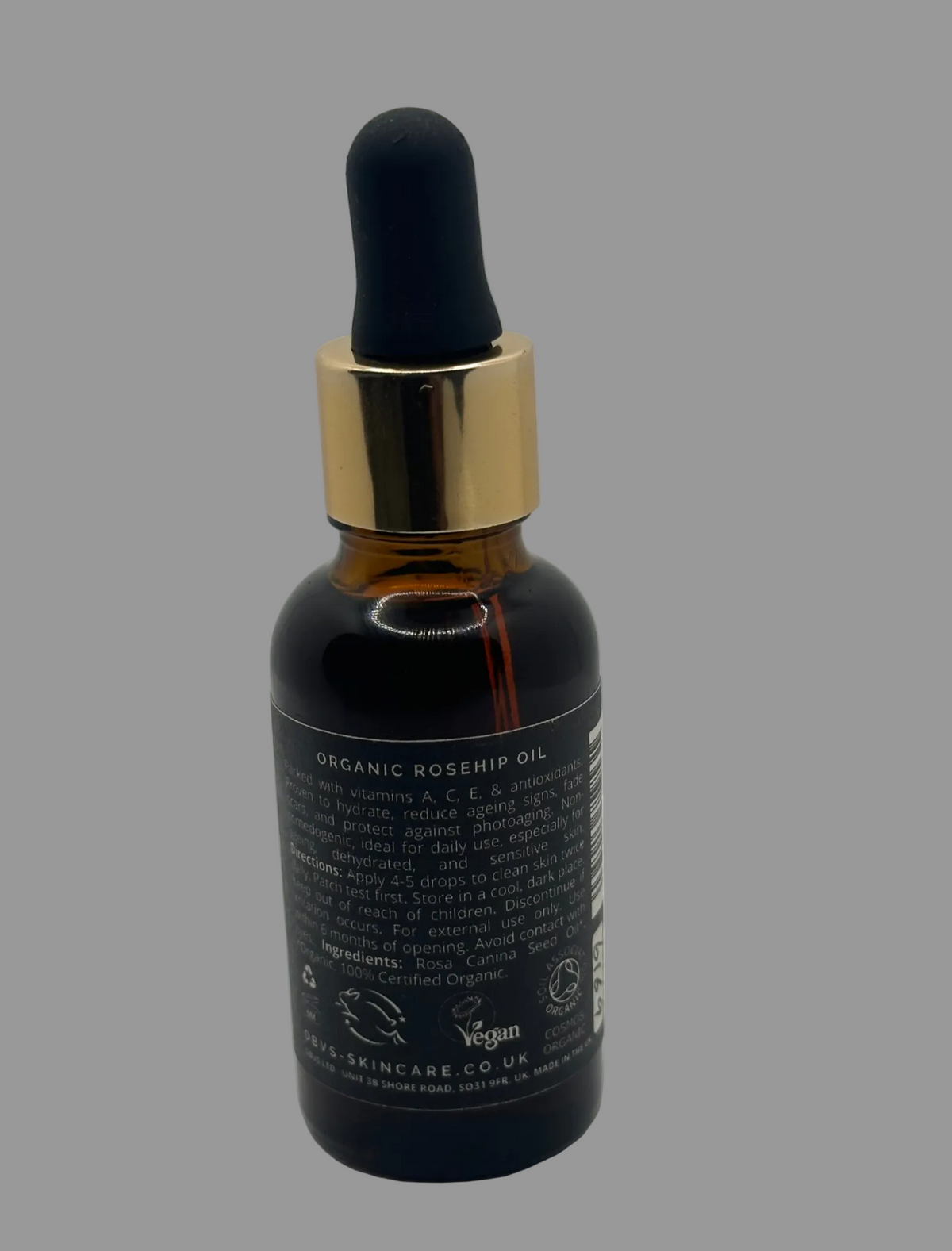 A dark amber glass dropper bottle of Obvs Rosehip Oil with a shiny gold collar and a black rubber bulb, shown against a neutral light gray background. Back of bottle shows that it contains vitamins A, C, E and antioxidents. It is certified Vegan, Leaping bunny and COSMOS Organic.