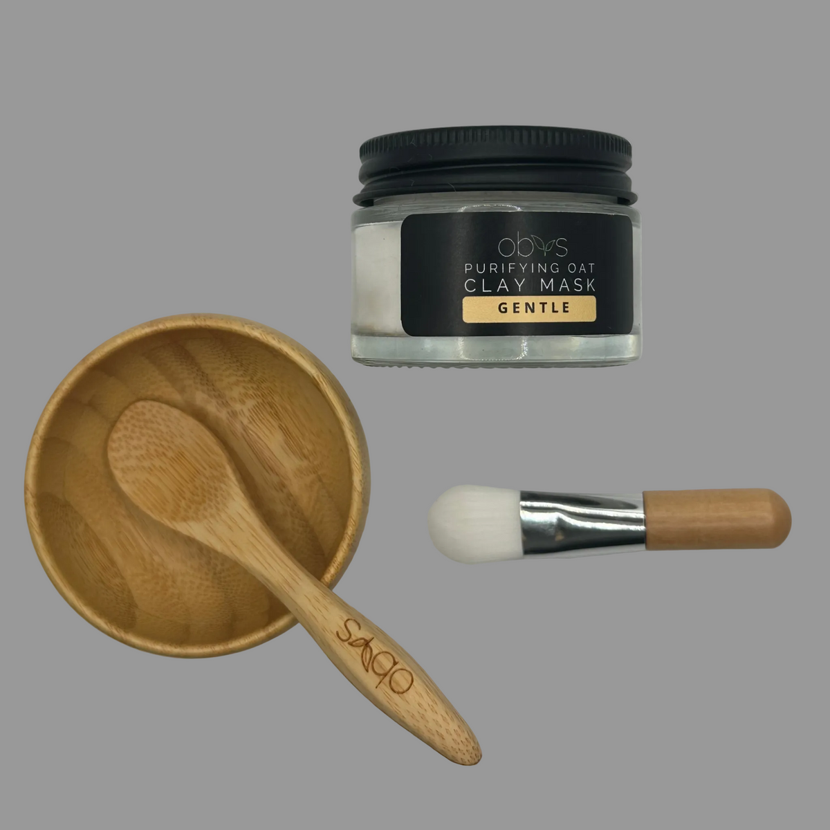 Obvs Face Mast Accessories - wooden spoon, bowl, soft white brush, lay on a slighly textured white cloth.  They lay next to the Obvs Purifying Oat Clay Mask Gentle (sold separately).